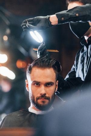 Front view of young bearded man that sitting and getting haircut in barber shop by guy in black protective maskの写真素材