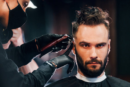 Front view of young bearded man that sitting and getting haircut in barber shop by guy in black protective maskの写真素材