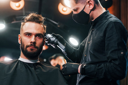 Front view of young bearded man that sitting and getting haircut in barber shop by guy in black protective maskの写真素材