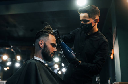 Young bearded man sitting and getting haircut in barber shop by guy in black protective maskの写真素材