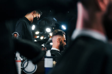 Young bearded man sitting and getting haircut in barber shop by guy in black protective maskの写真素材