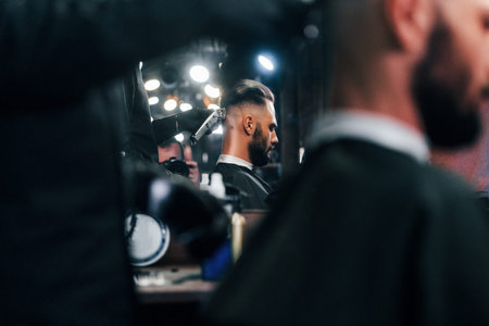 Young bearded man sitting and getting haircut in barber shop