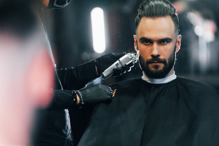Young man with stylish hairstyle sitting and getting his beard shaved in barber shopの写真素材