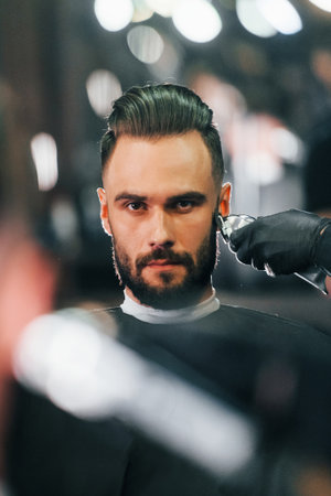 Young man with stylish hairstyle sitting and getting his beard shaved in barber shopの写真素材