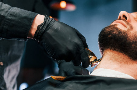 Young man with stylish hairstyle sitting and getting his beard shaved in barber shopの写真素材