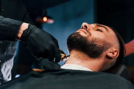 Young man with stylish hairstyle sitting and getting his beard shaved in barber shopの写真素材