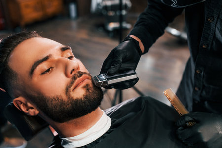Young man with stylish hairstyle sitting and getting his beard shaved in barber shopの写真素材