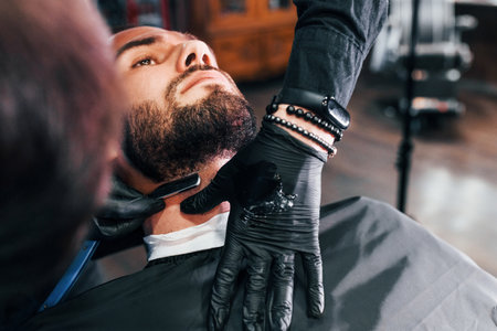 Young man with stylish hairstyle sitting and getting his beard shaved in barber shopの写真素材