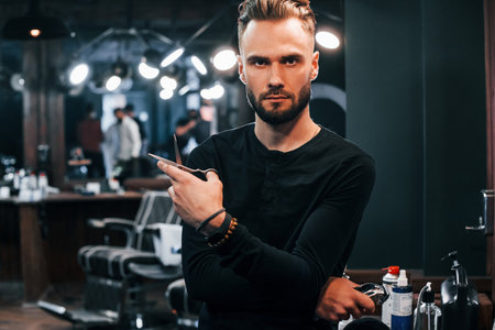 Young bearded man standing in barber shop and holding scissorsの写真素材
