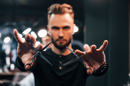 Young bearded man standing in barber shop and holding scissorsの写真素材