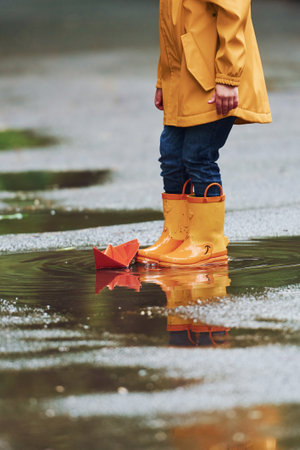 Kid in yellow waterproof cloak and boots playing with paper handmade boat toy outdoors after the rainの写真素材