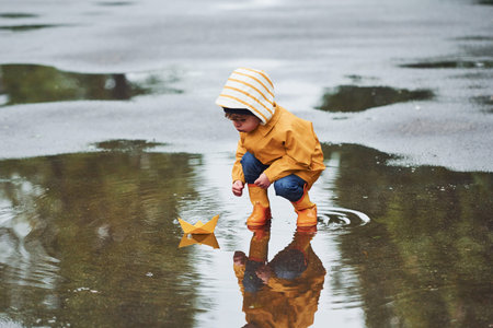 Kid in yellow waterproof cloak and boots playing with paper handmade boat toy outdoors after the rainの写真素材