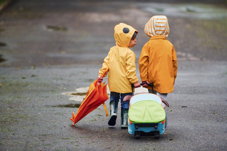 Two kids with umbrella, suitcase and yellow waterproof cloaks and boots walking outdoors after the rain togetherの写真素材