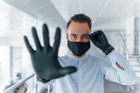 Showing stop gesture by hand. In protective mask and gloves. Young handsome man in formal clothes indoors in the office at daytimeの写真素材