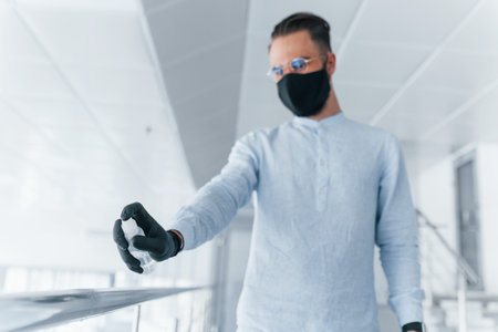 Holding antiseptic. In protective mask and gloves. Young handsome man in formal clothes indoors in the office at daytimeの写真素材