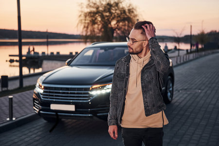 Handsome unshaved man in fashionable clothes walks near his black car at evening time