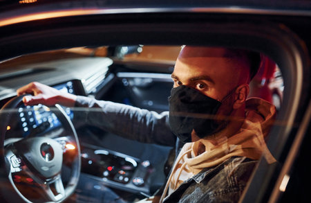 In black protective mask. Handsome unshaved man in fashionable clothes riding his automobileの写真素材