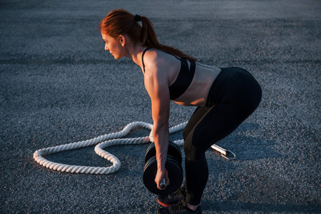 Lifting weights. Woman in sportswear have fitness day on the road at evening timeの写真素材