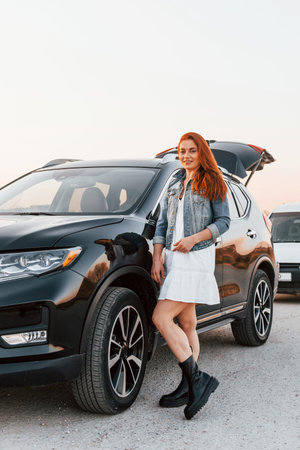 Redhead woman in skirt standing near black automobile on the road