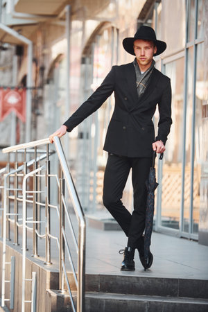 Elegant young man in black hat and with umbrella outdoors in the cityの写真素材