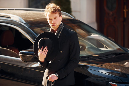 Standing near black car. Elegant young man in formal classy clothes outdoors in the cityの写真素材