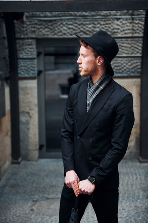 Elegant young man in black hat and clothes with umbrella in hands outdoors in the cityの写真素材
