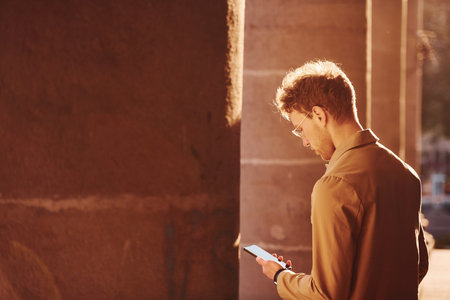 Nice sunshine. Holding phone in hand. Elegant young man in formal classy clothes outdoors in the cityの写真素材
