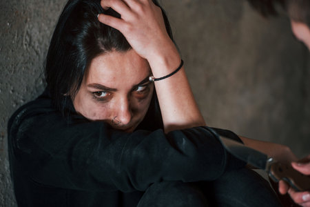 Kidnapped woman sitting and threatened by man with knife in hands in abandoned buildingの写真素材