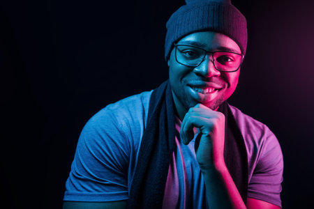 In hat and scarf. Futuristic neon lighting. Young african american man in the studioの写真素材