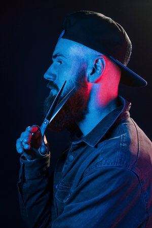 With scissors. Young bearded barber in cap standing in the studio with neon lightingの写真素材