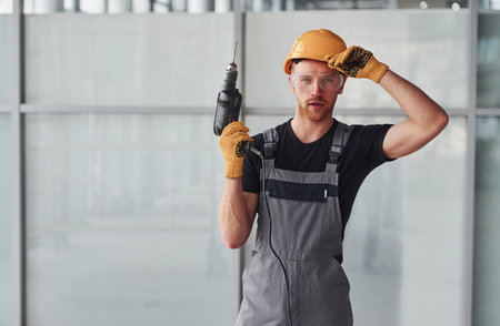 Man in grey uniform with drill in hand standing indoors in modern big office at daytimeの写真素材
