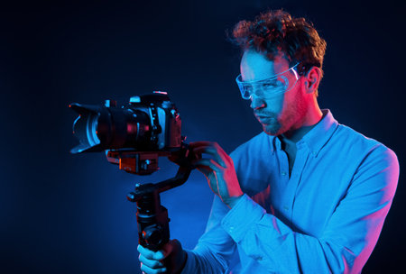 Holds camera. Neon lighting. Young european man is in the dark studioの写真素材
