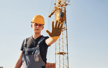 Shows stop sign by hand. Construction worker in uniform and safety equipment have job on buildingの写真素材