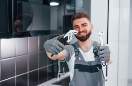 Young professional plumber in grey uniform standing on the kitchen and holding work equipment in...