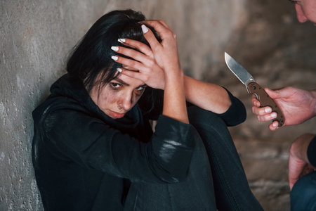 Kidnapped woman sitting and threatened by man with knife in hands in abandoned buildingの写真素材