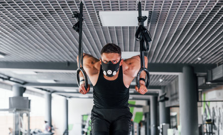 Exercising on rings. Young sportive strong man in black wear and mask have workout day in gymの写真素材