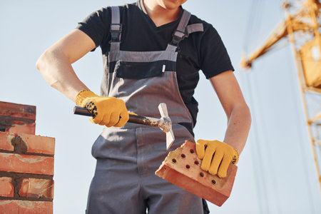 Holding brick and using hammer. Construction worker in uniform and safety equipment have job on buildingの写真素材