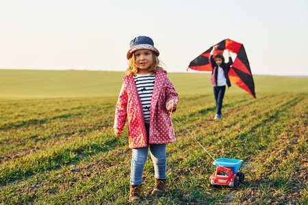 Two little girls friends have fun together with kite and toy car on the field at sunny daytimeの写真素材