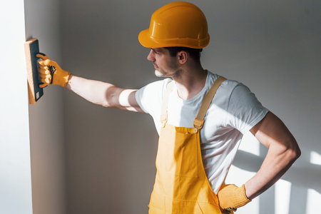Handyman in yellow uniform polishing wall indoors. House renovation conceptionの写真素材