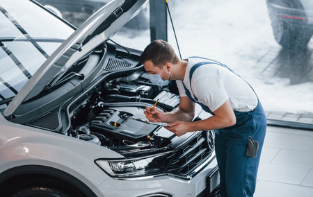 Young man in white shirt and blue uniform repairs automobileの写真素材