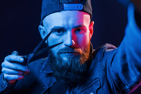 With scissors. Young bearded barber in cap standing in the studio with neon lightingの写真素材