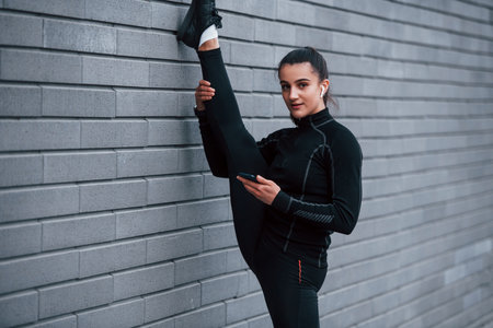 Young sportive girl with phone in black sportswear doing legs stretching outdoors by using gray wallの写真素材