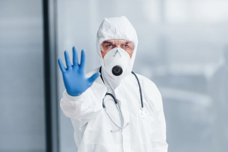 Shows stop gesture by hand. Male doctor scientist in lab coat, defensive eyewear and maskの写真素材