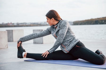 Young sportive girl doing yoga exercises on fitness mat outdoors near lake at daytime