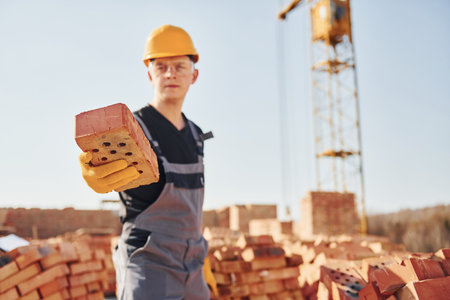 Holds brick in hand. Construction worker in uniform and safety equipment have job on buildingの写真素材