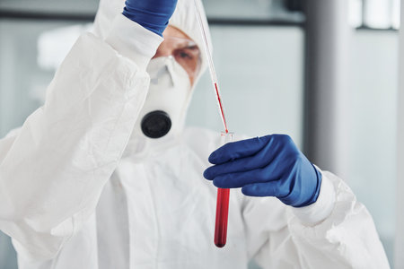 Male doctor scientist in lab coat, defensive eyewear and mask works with test tube with blood inside of itの写真素材