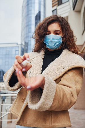 Portrait of young girl with curly hair in protective mask that using antiseptic outdoors near business building at quarantine time. Conception of coronavirusの写真素材