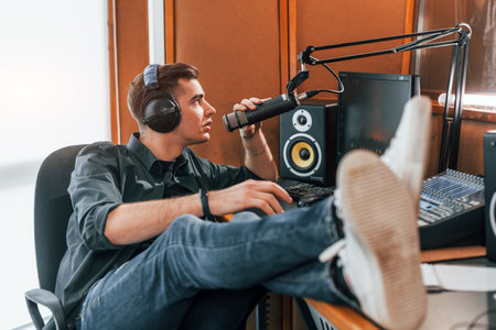 Talks and uses mic. Young man is indoors in the radio studio is busy by broadcastの写真素材