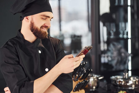 Professional young chef cook in uniform standing with phone on the kitchenの写真素材