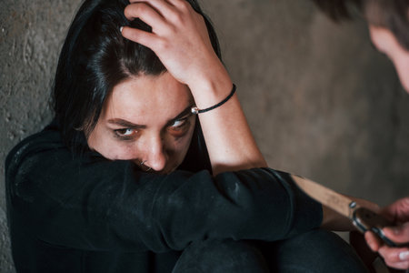 Kidnapped woman sitting and threatened by man with knife in hands in abandoned buildingの写真素材